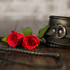 A red collar and two red roses on a wooden table. There is also a black whip in the image.
